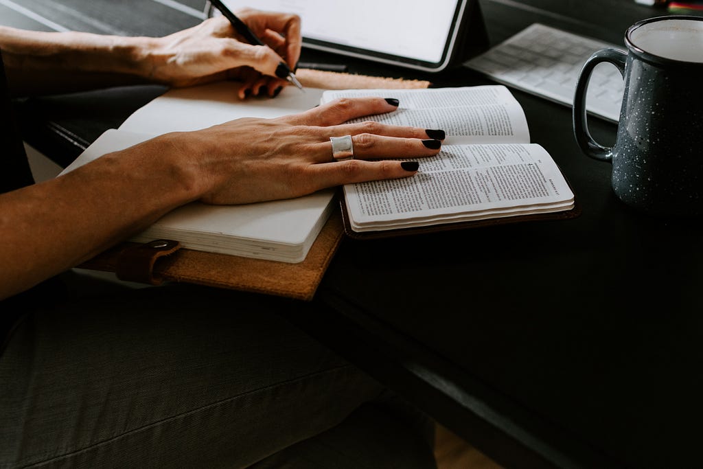 A woman’s hand holding a book open and writing in a journal.