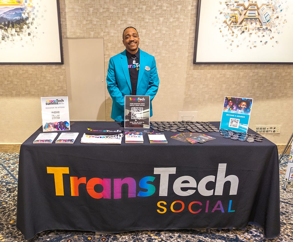 E.C. Pizarro III, a Black man wearing a teal jacket, smiles while standing behind a table with fliers, buttons, and a black drape reading “TransTech Social” in rainbow-colored lettering.