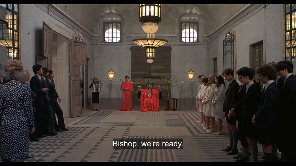 A wide white room with the captives gathered at either side and the Bishop at the far end in the middle, extravagantly dressed to officiate the ‘wedding.’ Again, we see a chandelier, wide doors, windows, and mirrors with elaborate framing. The captives have been dressed in suits and dresses. Subtitles on the screen read “Bishop, we’re ready.”