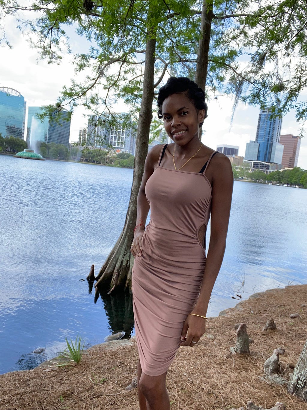 Photo of the author posed with a hand on her hip in a beige dress smiling and standing near the lake in Downtown Lake Eola.