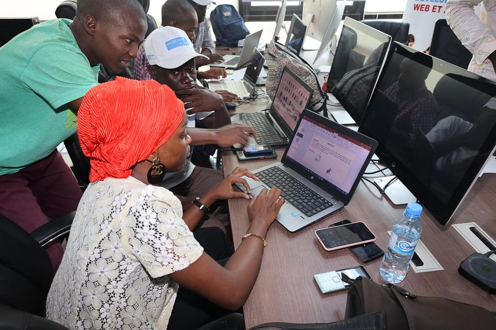 Three participants at a hackathon are discussing with one another. Two are seated in front of their laptop computers, with one participant standing further back as if providing directions. The participant seated on the foreground is a woman wearing a white embroidered shirt and an orange headdress. The participant who is standing is wearing a light green shirt. The other participant who is seated is wearing a white baseball cap with light blue lettering.