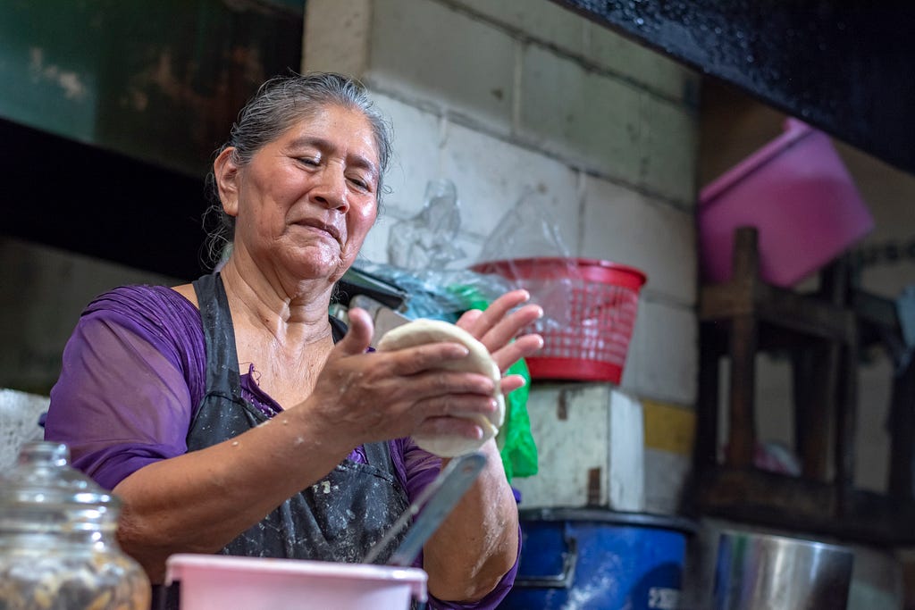 Woman in an apron shaping dough in her hands in a small store with concrete walls.