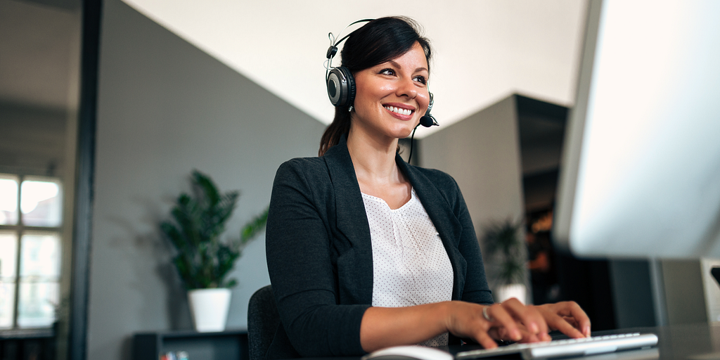 A smiling woman with a headset types at her computer.