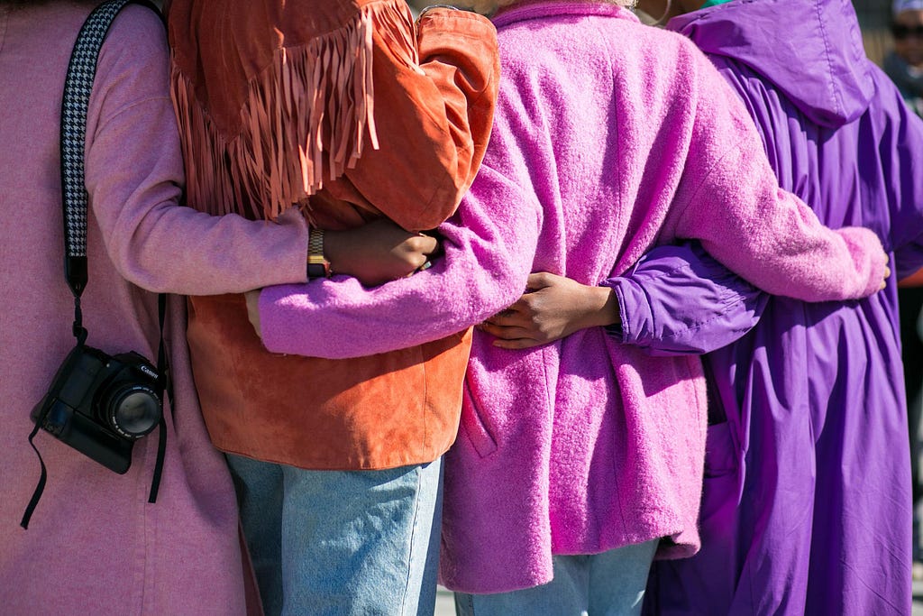Four women hugging. Image taken from behind showing their backs and arms.