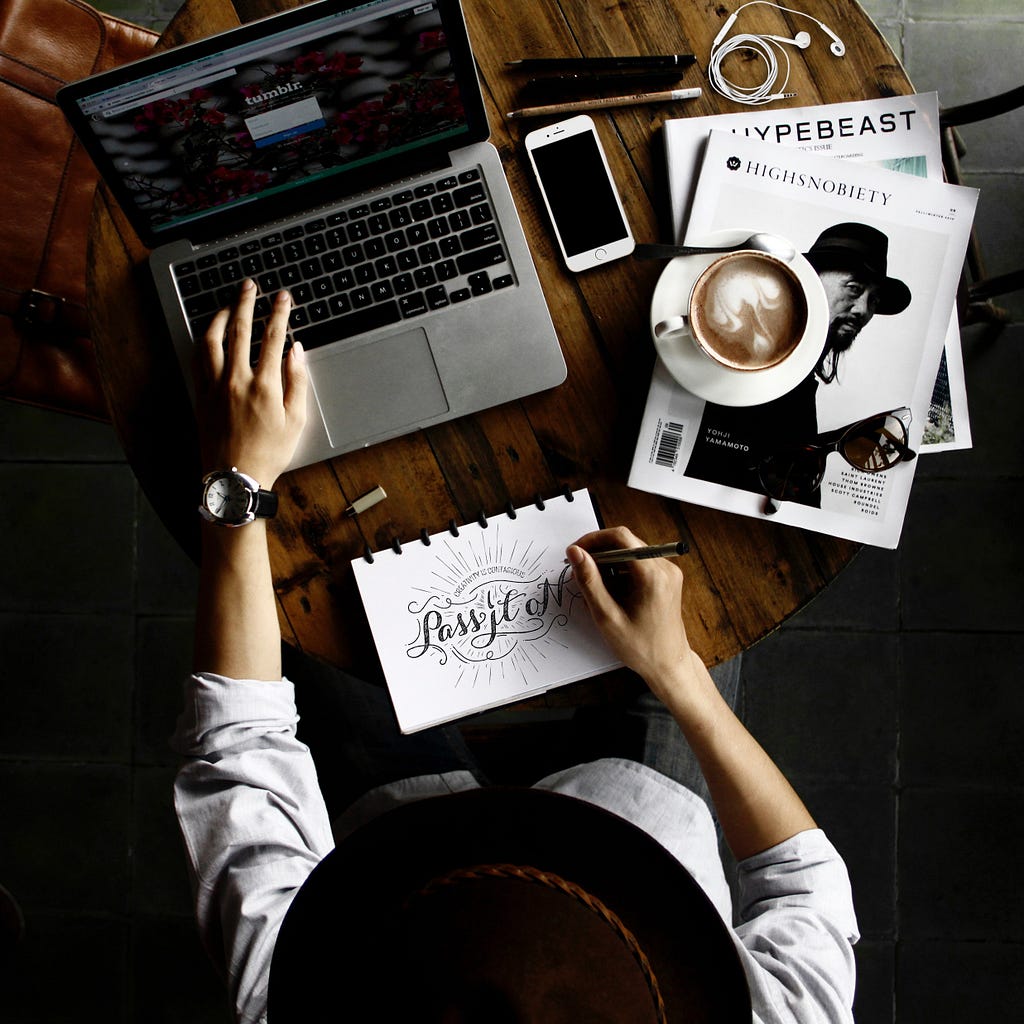 Person with computer, a sketchpad with words “Pass it on”, latte, iPhone, earphones, and magazines