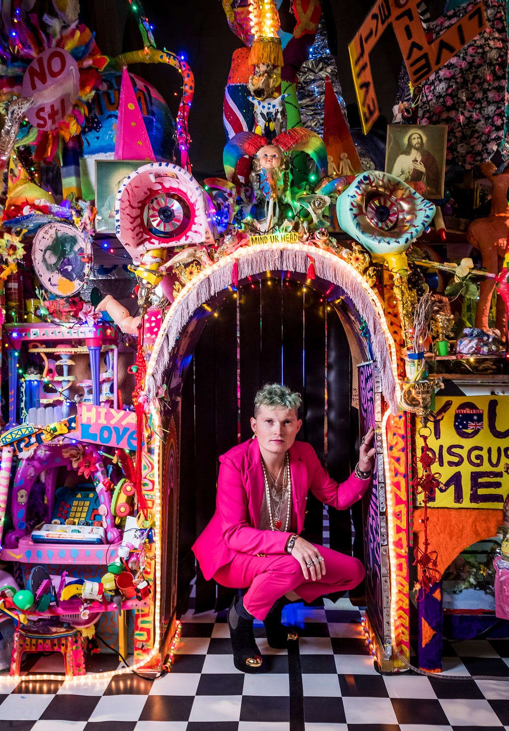 Paul Yore crouches in the archway of his installation ‘World made flesh’. Technicolor ephemera covers the walls, including hand painted posters with phrases such as ‘hiya love’ and ‘you disgust me’.