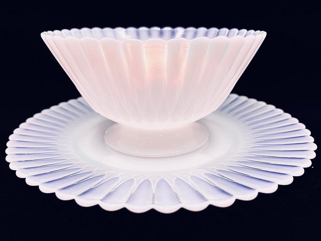 A full-frontal view of a Monax Petalware compote dish on top of a matching dessert plate against a black backdrop. Both the border of the plate and the scalloped lip of the dish in the background are translucent here, while the scalloped lip of the dish in the foreground seems lit from within, with opalescent fire.