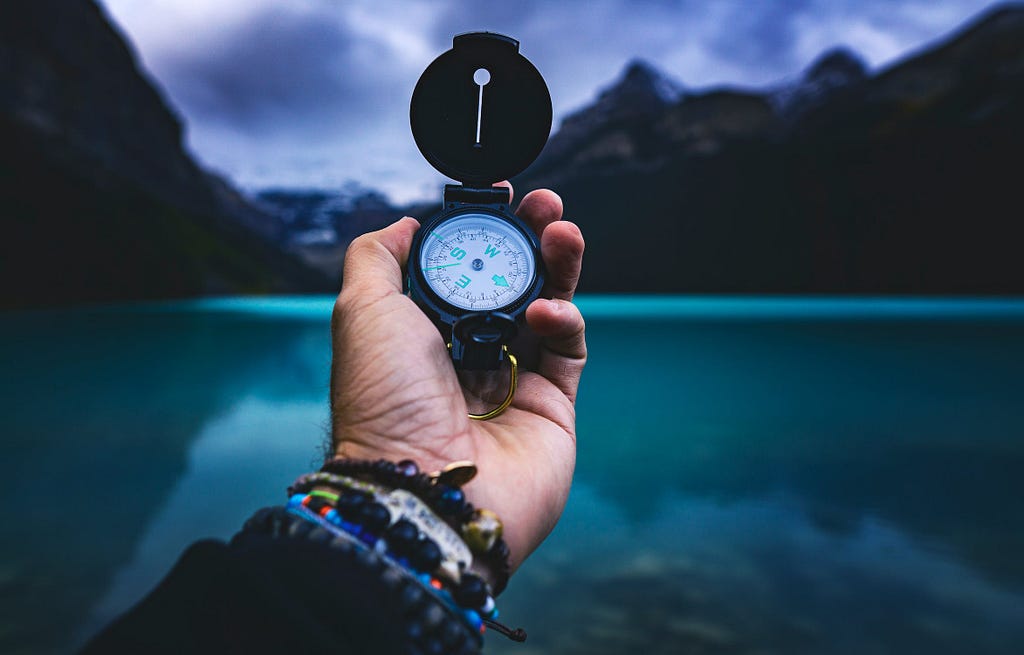 Person Holding A Compass. In the background, it’s clean green colored water and mighty mountains with their shadows in the water. It’s beautiful.
