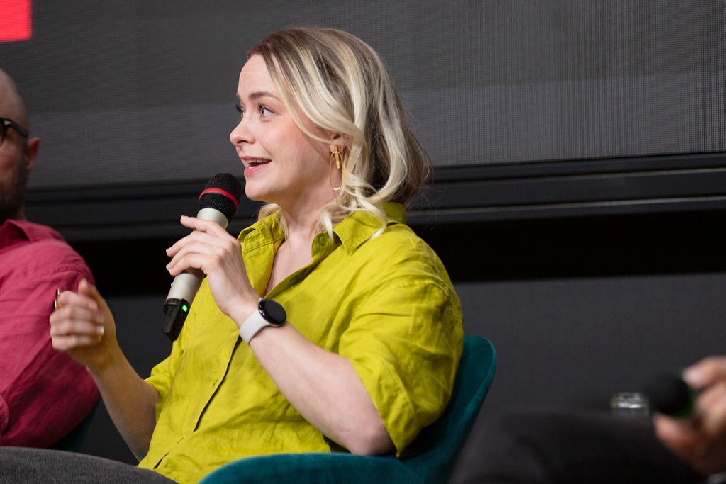 A person with blonde hair and a bright chartreuse shirt, seated in a chair and speaking into a microphone.