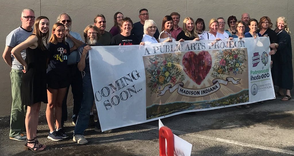 A large group of community members smiles and stands behind a sign announcing, “Coming soon… A public arts project!”