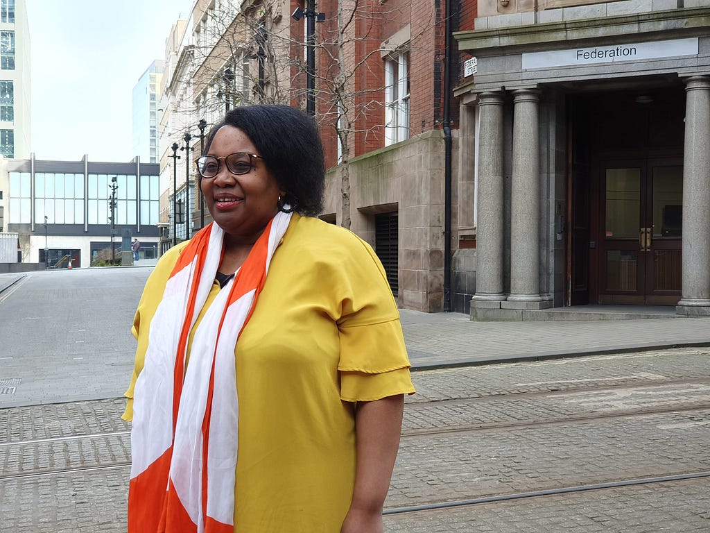 Reina is wearing a bright yellow shirt and orange and white scarf and is looking away from the camera. She is standing across the road from Federation House in Manchester.