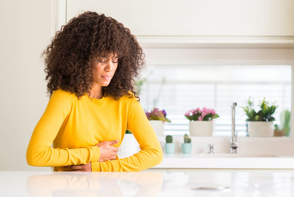 A Black woman with natural hair, wearing a yellow sweater and standing in a kitchen, holds her stomach as if nauseous