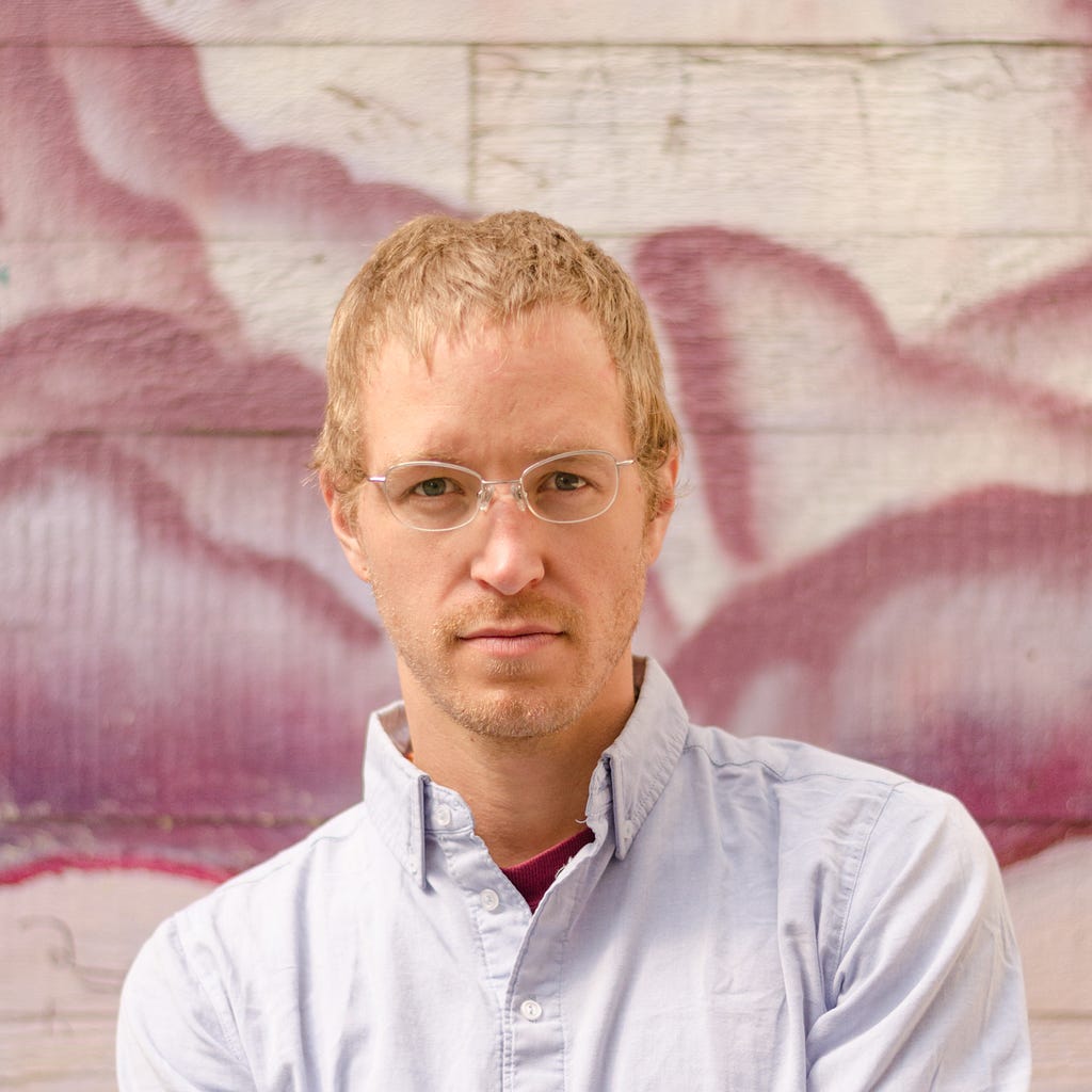 Lex Pelger, a white man in his thirties with short blond hair and glasses. He is wearing a white shirt and standing in front of a white wall painted with lilac clouds.