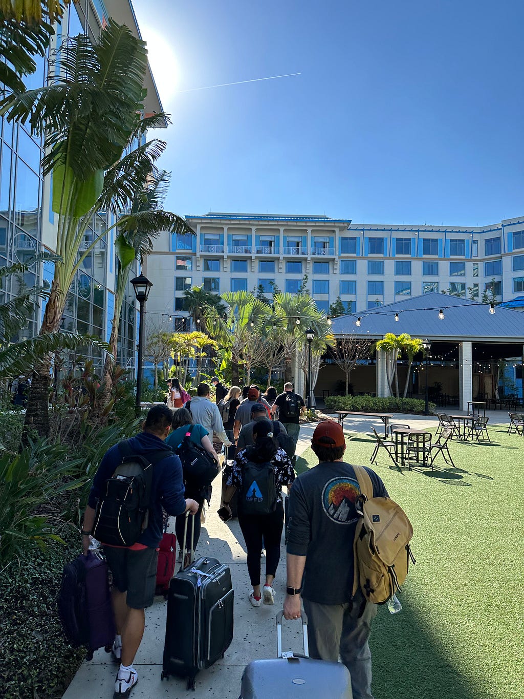A group of people with suitcases in hand walk toward a hotel passing by a courtyard.