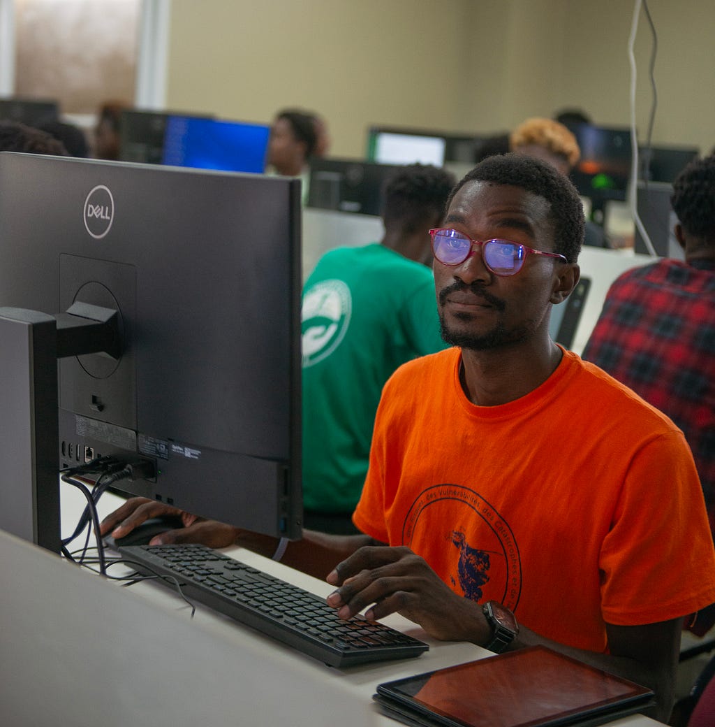 A student in a bright orange T-shirt looks intently at a computer screen as the reflection from the monitor appears on his glasses.