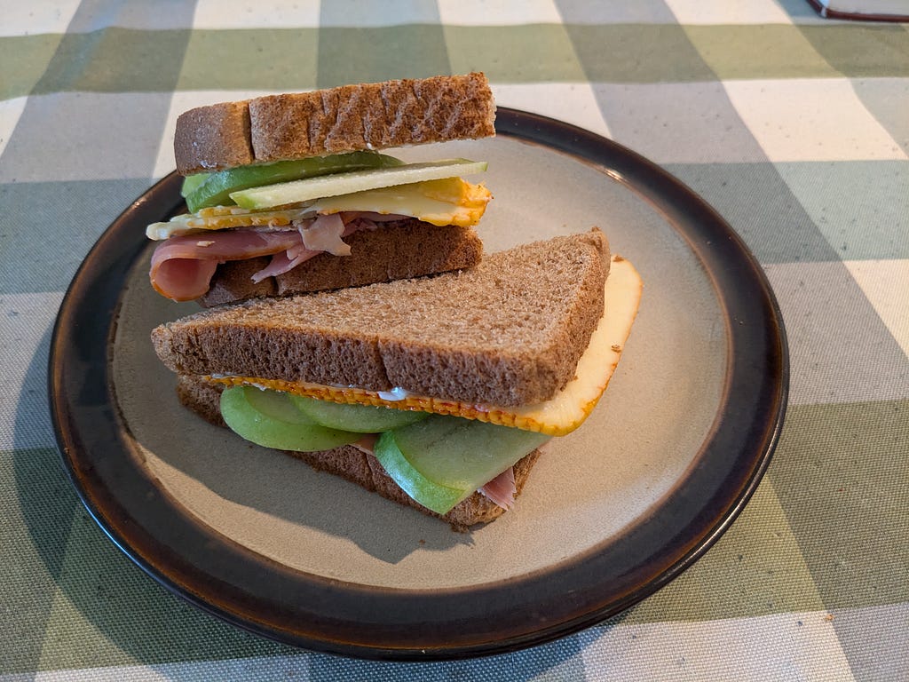 Ham, muenster and granny smith apple sandwich on whole wheat bread, a.k.a. “toast,” served on a beige plate with a brown rim, sitting on a green, blue, and white gingham tablecloth.