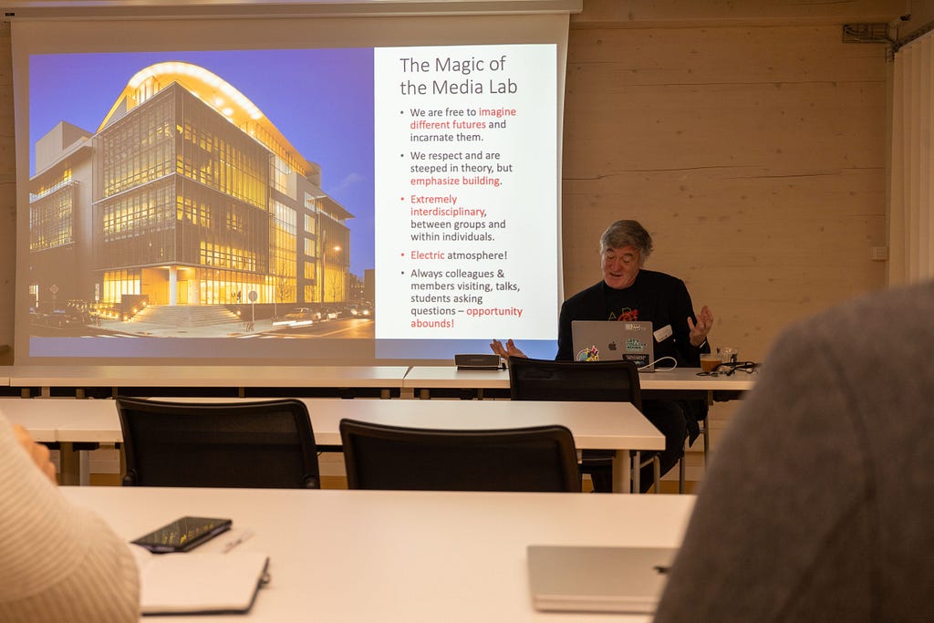 Joe Paradiso wears a black jacket and sits at a lecture hall table, with a laptop on the table in front of him, presenting slides displayed on a projector screen behind him; the screen displays an image of the Media Lab building E14 lit up with yellow/gold light on a dark blue evening sky backdrop.