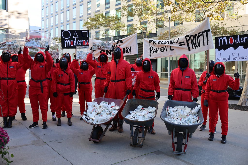 A group of people wearing red jumpsuits with black belts push three wheelbarrows and hold aloft clear boxes full of fake money. The people all have their faces covered with a black face covering so that no part of their face is visible, and on the covering is either a white square, triangle or circle. Members of the crowd hold up signs that read “Prolonged pandemic = Profit” and the three shapes and with “Moderna.” The costumes and shapes are reference to the Netflix series, “Squid Game.”