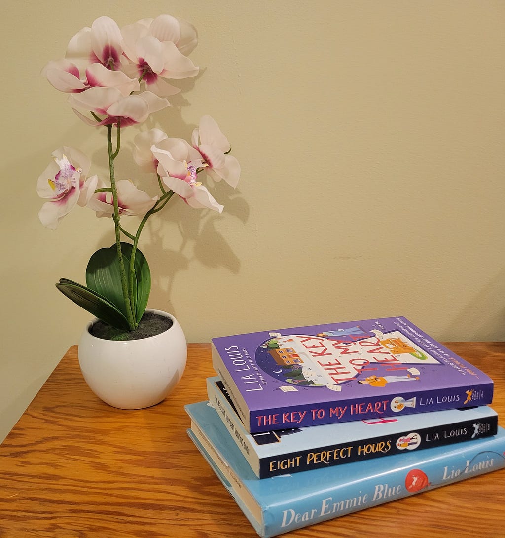 A stack of books by Lia Louis placed on a wooden table beside a pot of orchids