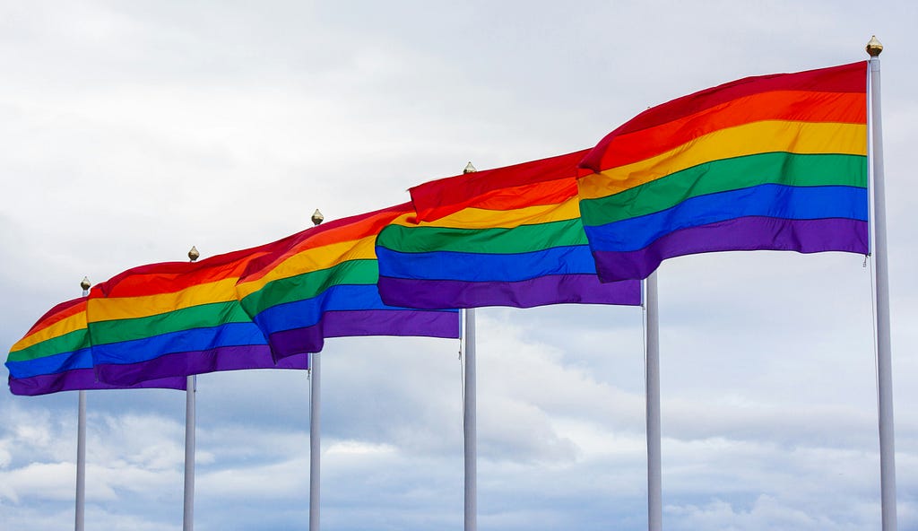 A photo of six rainbow flags flying on flag posts, with clouds in the background. The flag colours from the top down are dark red, orange, yellow, green, blue and purple.