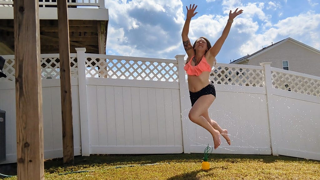 Female jumping over a small pineapple sprinkler in a backyard. Behind her is a white privacy fence. The yard is yellow-green. The female is in a pink top and black bottoms. Her arms are spread above her head, fingers spread, her face has a big smile and her feet are off the ground, in the air.