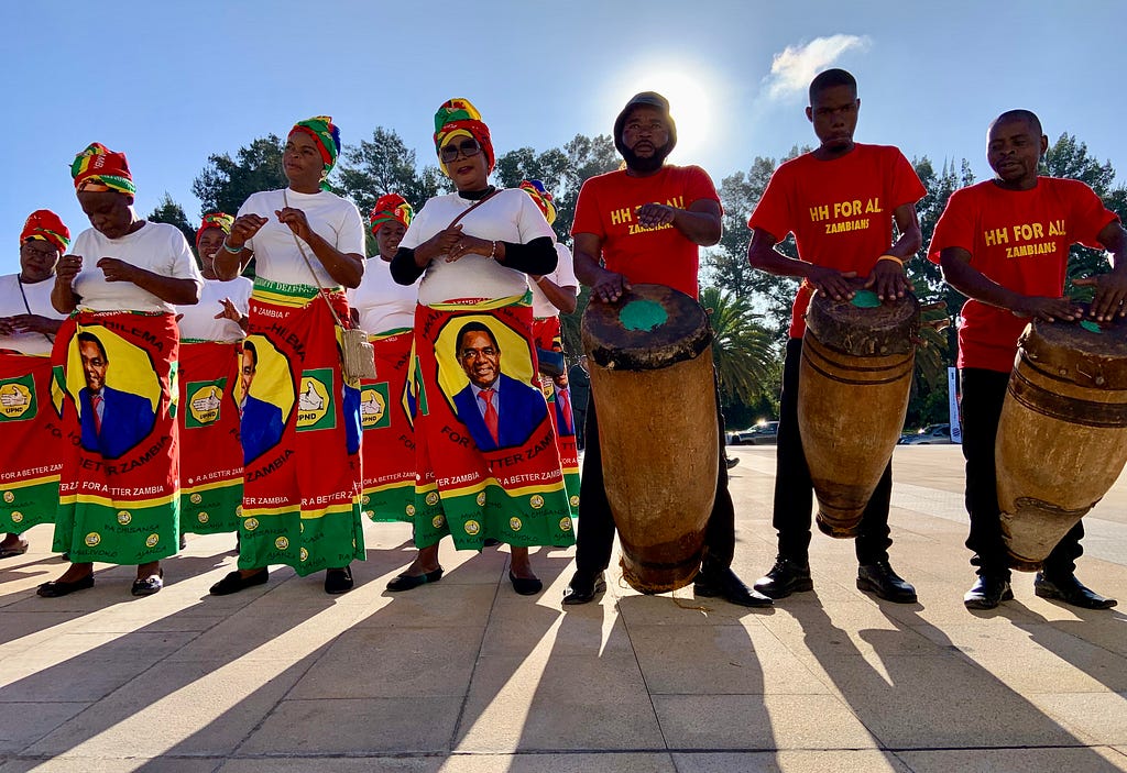 Colorfully dressed dancers and drummers perform at the opening of Summit for Democracy in Zambia as the sun behind them casts shadows of them all.