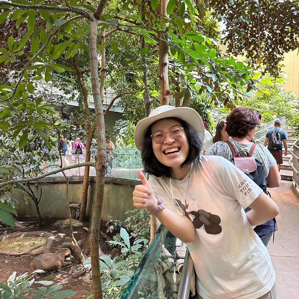 A Queer Asian non-binary person wearing round glasses, a white hat, and T-shirt smiles and gives a thumbs-up at a zoo with a capybara nearby.