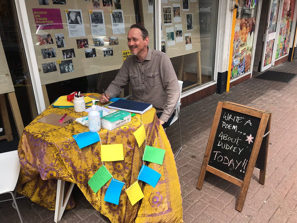 A photograph of local poet Rick sitting at a table smiling. A blackboard next to him says “write a poem… about Dudley TODAY!” The table Rick is sitting at is on the pavement of Dudley High Street. It is covered in a gold and purple coloured fabric. On the table are pens, paper, hand sanitiser and a box of face masks. A number of A5 coloured pieces of paper are hanging down the side of the table, strung together to make a poetry paperchain.