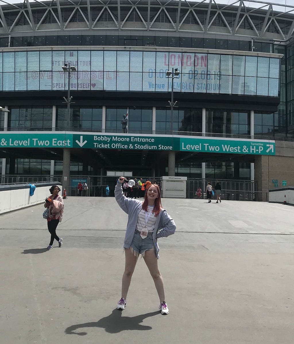 Author (female, white, 21) stands in front of Wembley Stadium in denim shorts and a grey hoodie holding an ARMY Bomb.