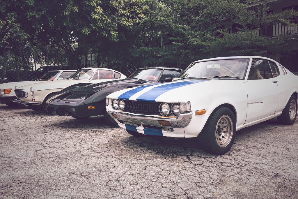 Image of a lineup of classic cars parked outdoors, featuring various models, including a white car with a blue racing stripe in the foreground.