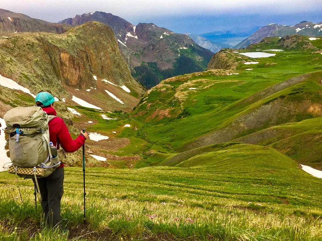 A man with a large backpack and walking sticks looks at a green mountain valley with snowy mountain peaks in the distance