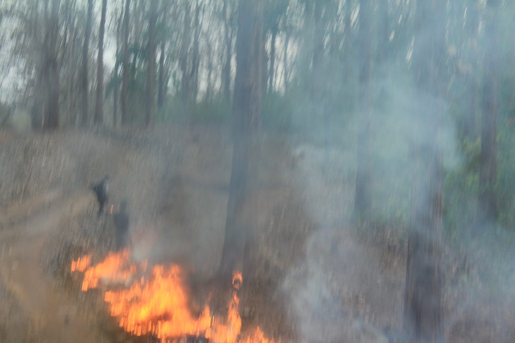 A blurred image of burning pyre and smoke. Some blurred human figures in the background.