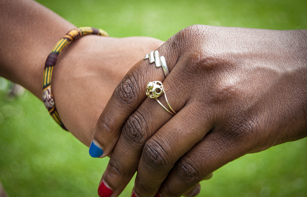 Two hands, Black and Brown, holding. One has rings and the fingernails painted in red and blue. The other one has a crafted bracelet in tones of brown, yellow and green.