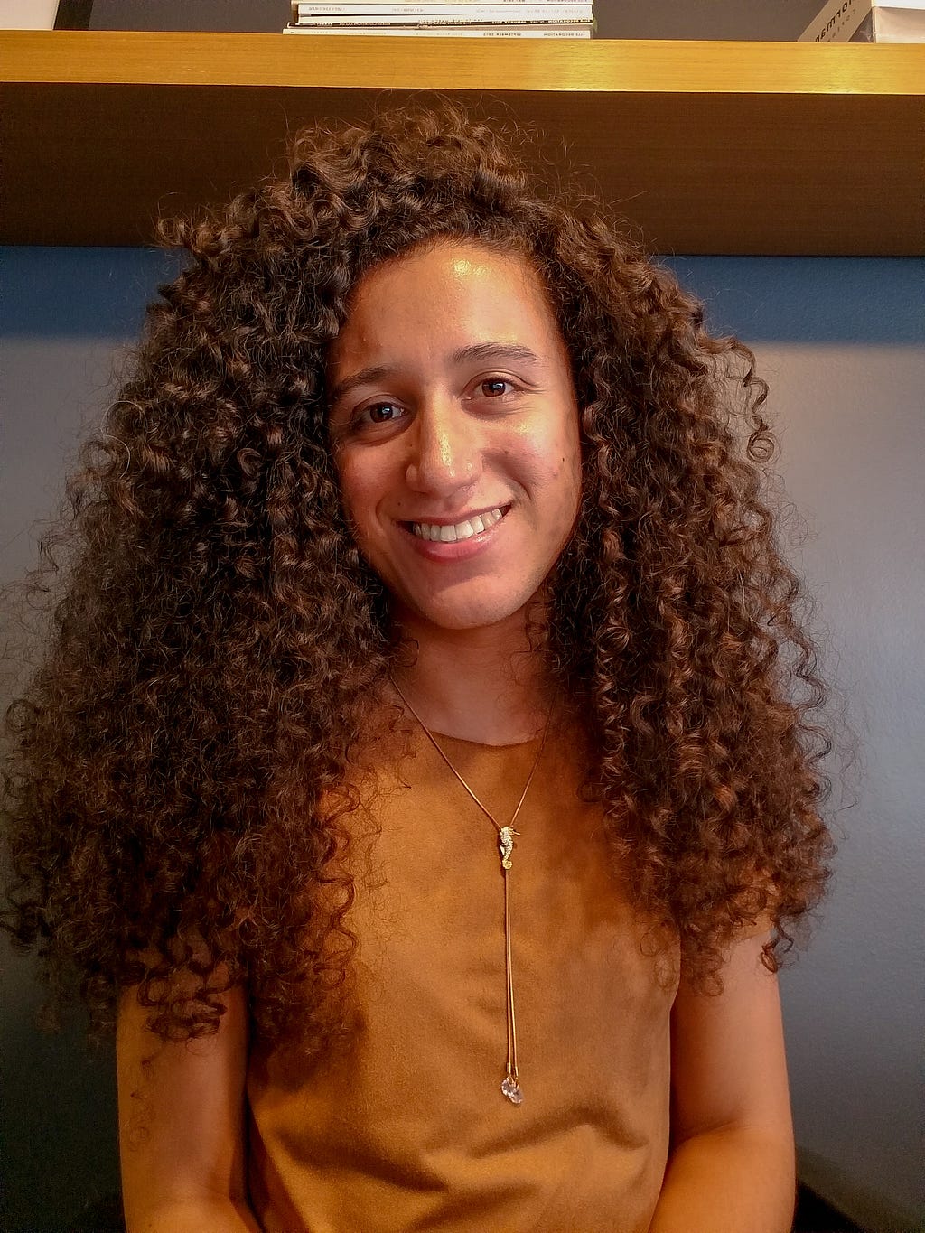 Jovem mulher negra e trans, de cabelo comprido e cacheado, sorrindo e usando blusa caramelo e um pingente dourado