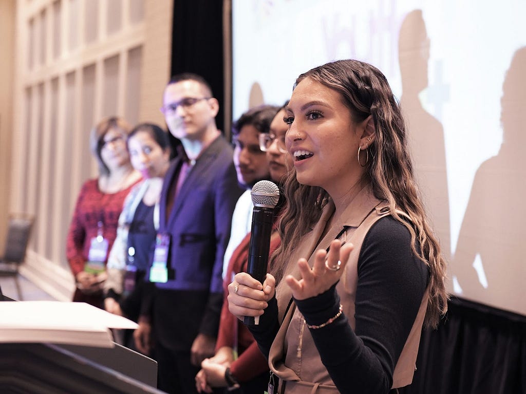 Melissa Holguín Pineda, NPNA’s Program Manager of the Inclusive Initiatives with a microphone in her hand, introducing NAYLC Cohort Leaders at NIIC 2022. The photo depicts New American Youth Leadership Council (NAYLC) participants at the Youth Leadership Summit at the National Immigrant Inclusion Conference in National Harbor, Maryland in December 2022.