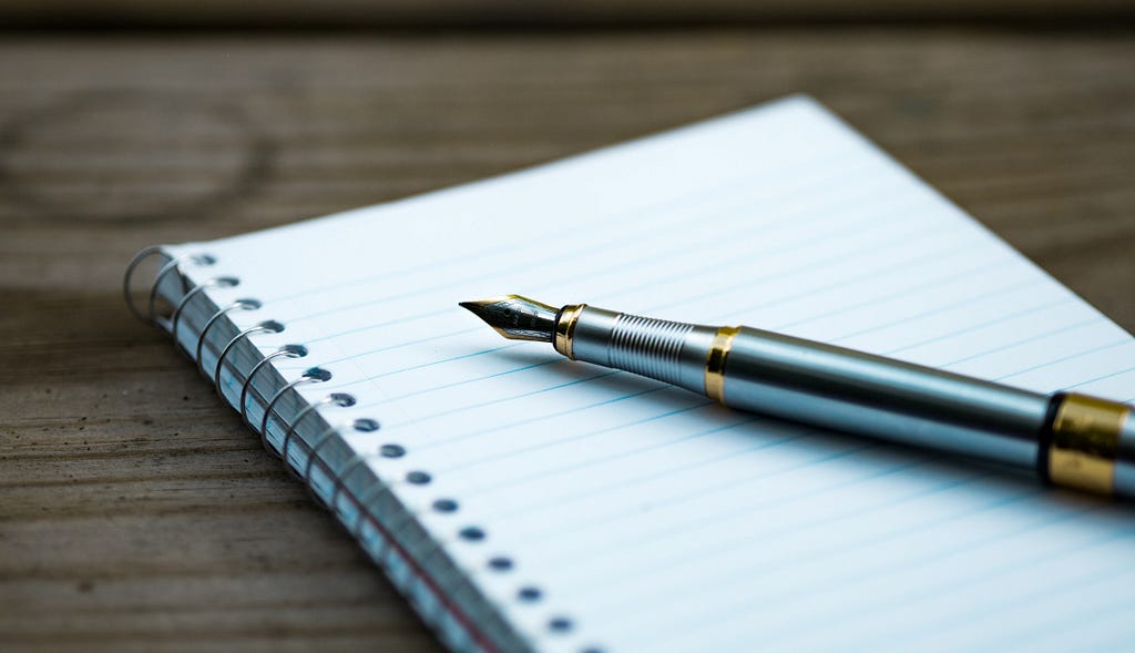 A fountain pen rests on an open, blank spiral notebook