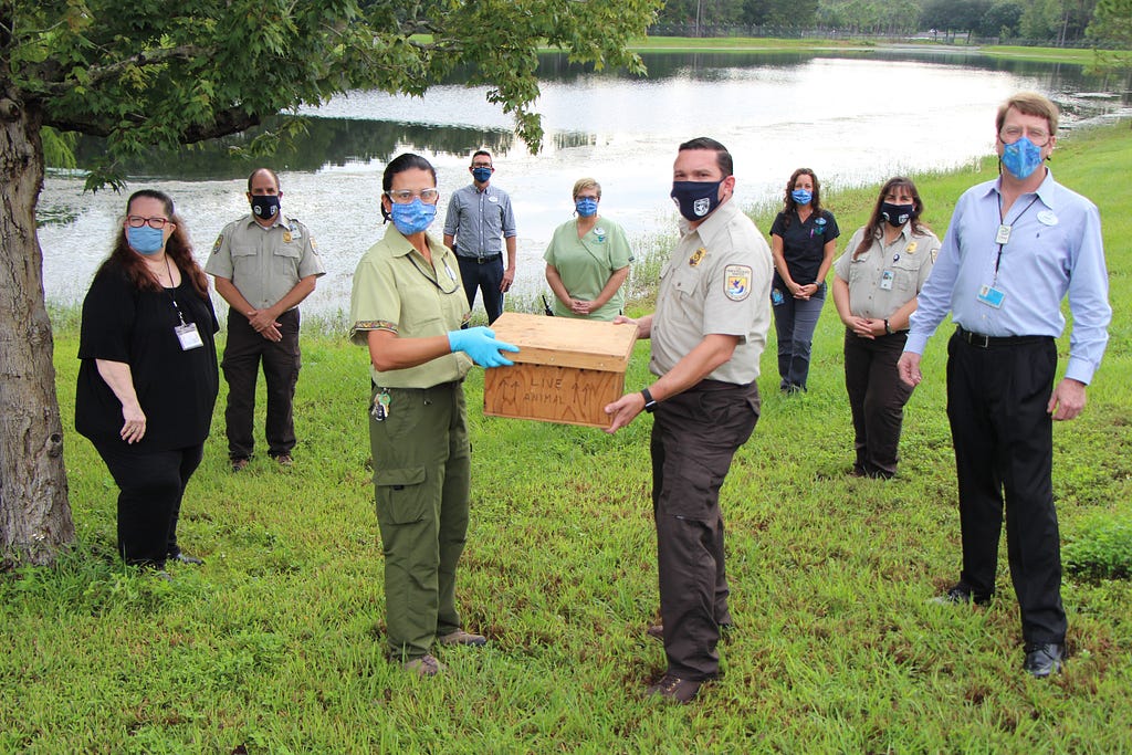 Group of people outside, all with face coverings. 2 hold wooden box