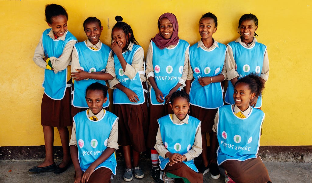 A group of girls in Splash vests smile and giggle in front of a yellow wall at school in Addis Ababa, Ethiopia.
