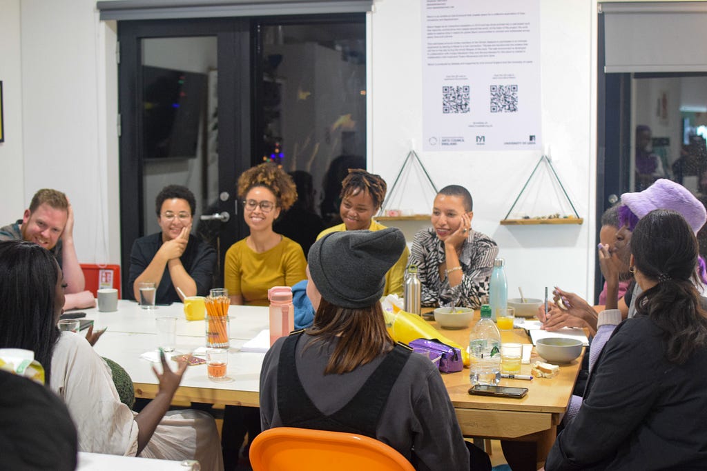 A group of people mid-smiles and laughter, engaged in conversation around a long table at YARD. In the background on the left-hand side, double doors show a dark sky outside. To the right of the doors, a poster with a QR code in on the walls. The poster is for Akeelah Bertram’s work, ‘Return’.