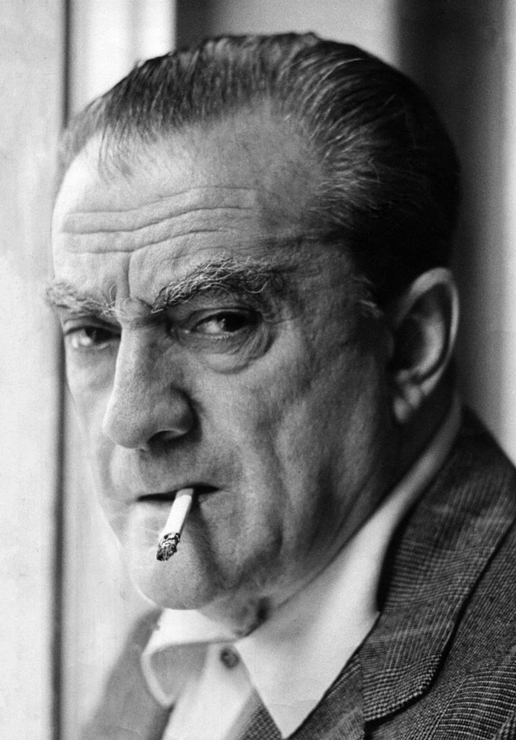 Black and white image of the face of Italian film director Luchino Visconti. Shot from the neck up as the old man glances over his left shoulder. He has wrinkled skin, a receding hairline, a serious expression, and a cigarette dangling from his lops.