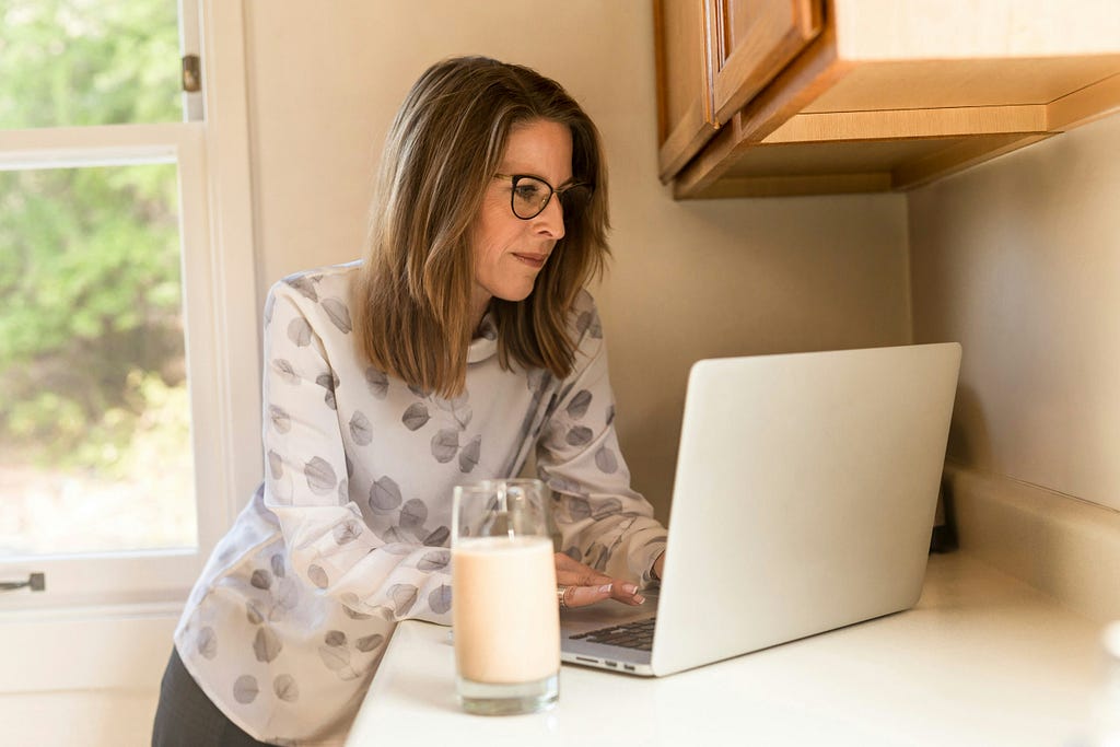 Woman stands at her laptop with a glass of milk.