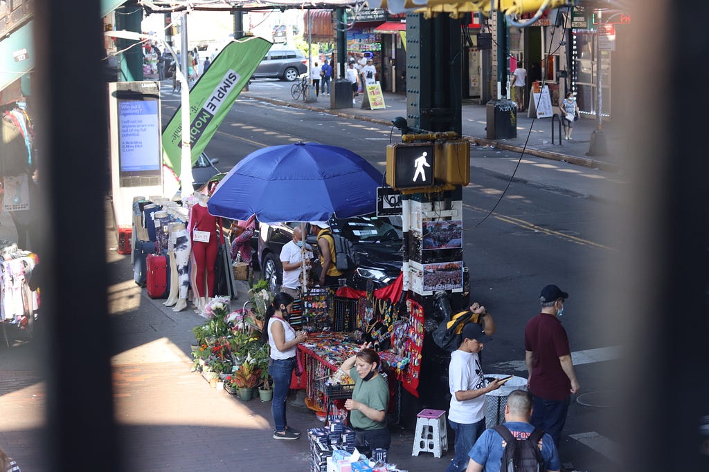 A photo of the corner of 82nd street and Roosevelt Avenue in Jackson Heights, Queens. There are individuals shopping from street vendors under a blue umbrella, a crossing street light, and people walking around