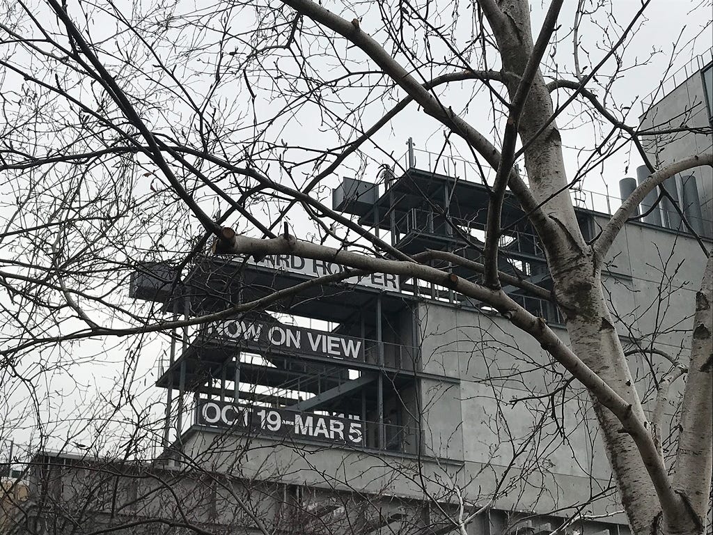 Bare trees, and a person atop a platform on a building. A sign on the building says Edward Hopper Now on View Oct 19-Mar 5.