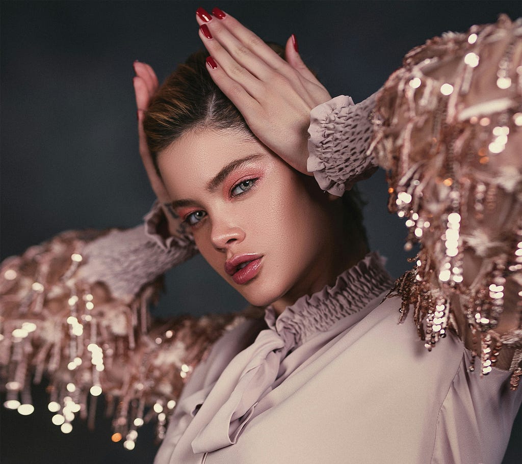 A young woman with glossy pink lips and shimmering eye makeup poses elegantly, wearing a blouse with sequin-embellished sleeves