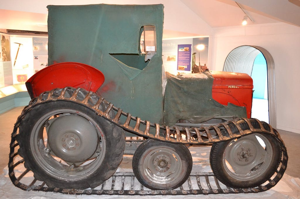 The converted ferguson tractor. An extra wheel is added. There are metal tracks covering the wheels. The body is painted bright red. Green canvas covers the vents and radiator. A green canvas box sits over the drivers seat tall enough for the driver to be able to stand up inside.