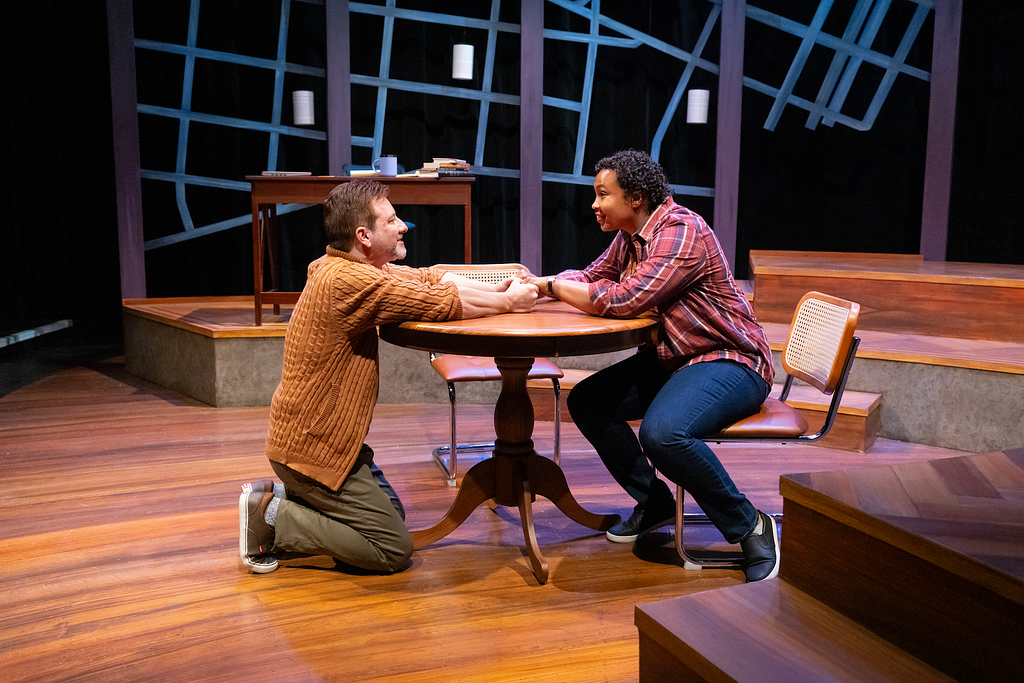 In a photograph from THE WANDERERS, a man in a cardigan kneels at a round table and smiles, holding the hands of a woman in a plaid shirt sitting at the table.