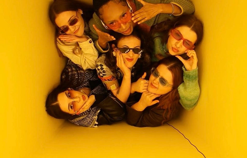 Six girls wearing sunglasses pose for a photo against a bright yellow background