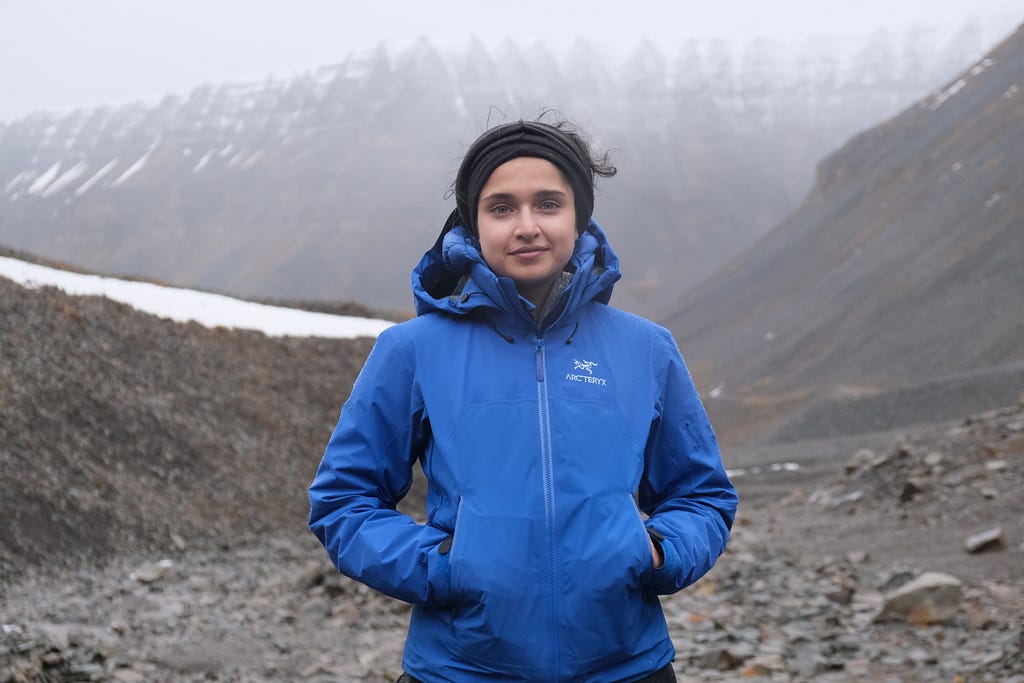 Sana Sharma facing the camera and wearing a blue winter jacket. Grey and brown, rocky and snow-dusted mountains in the background.