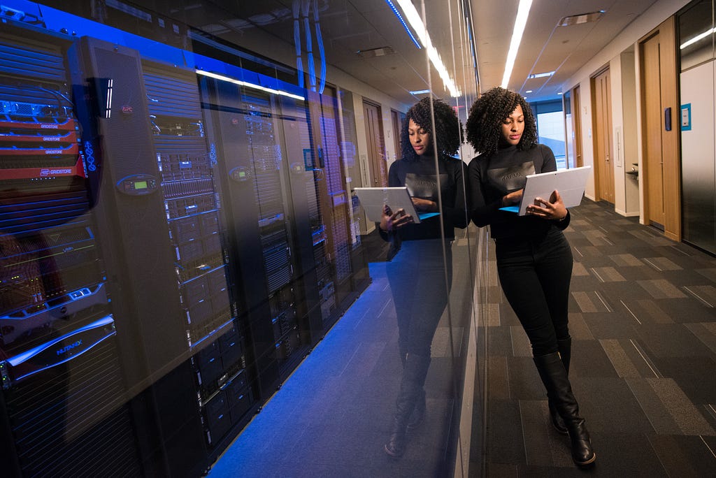 a woman holding a laptop in a hallway with doors on one side of her while she leans on a glass wall with racks of servers behind it.