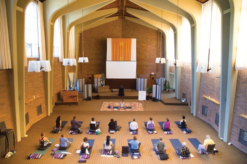 One of the large Yoga rooms at the Kripalu Center for Yoga and Health, showing rows of people on mats looking at an instructor at the center of the room, which resembles a high-ceilinged chapel.